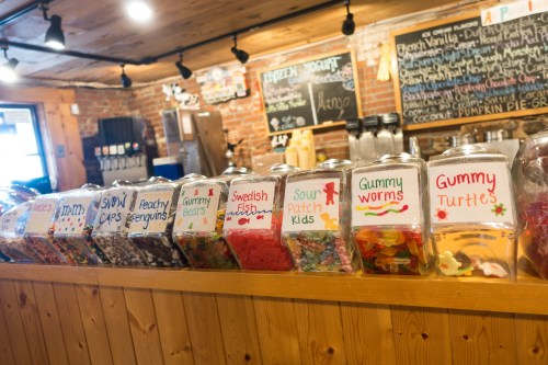 A variety of colorful candy jars lined up on a wooden counter, labeled with different candy names.