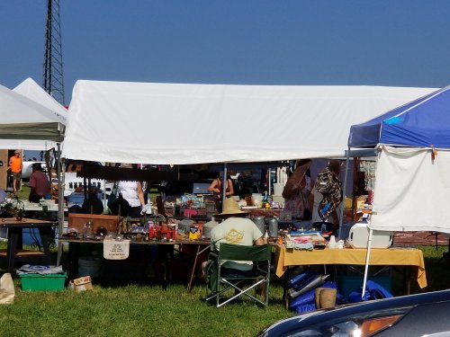A bustling outdoor market with various tents, tables filled with vintage items, and shoppers browsing under a clear blue sky.