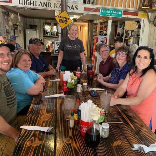 A group of eight people smiling around a wooden table in a restaurant, with a waitress standing in the center.