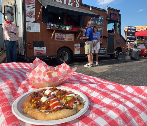 A food truck serves BBQ while a couple orders food at a fair, with a plate of loaded nachos in the foreground.