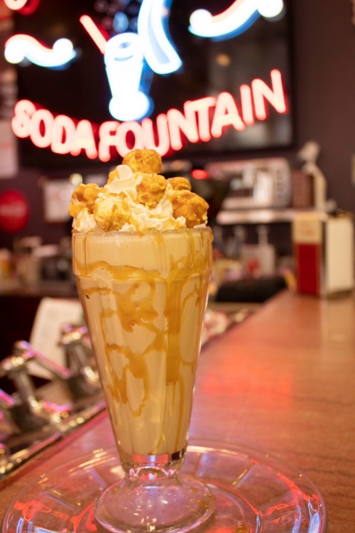 A tall glass of ice cream topped with whipped cream and caramel, garnished with popcorn, in front of a neon soda fountain sign.