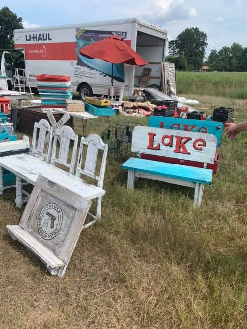 A grassy area with colorful wooden benches and signs, a U-Haul truck in the background, and various items for sale.