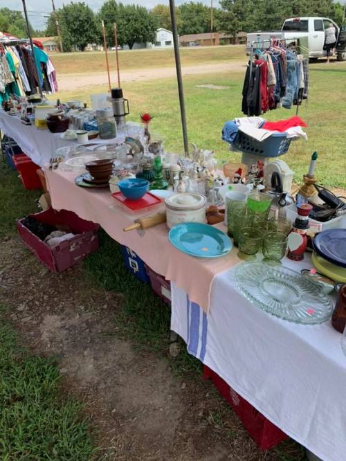 A table at a flea market displaying various household items, including dishes, glassware, and clothing.
