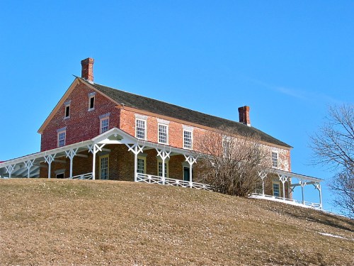 Historic brick house on a grassy hill, featuring a porch and blue sky in the background.