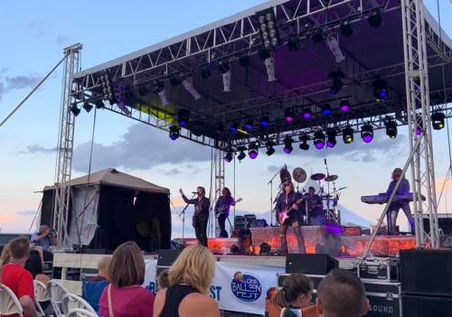 A live band performs on stage at an outdoor festival, with a colorful sunset in the background and an audience in front.