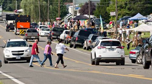 A busy street scene with people crossing, parked cars, and vendors set up along the roadside during an event.
