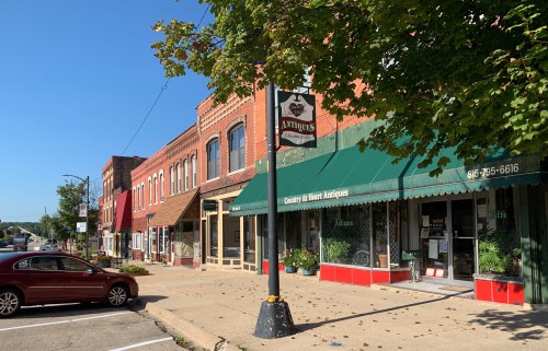 A quaint street lined with brick buildings and shops, featuring an antique store with a green awning on a sunny day.