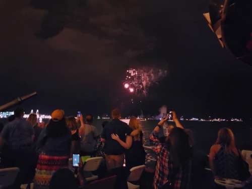 People on a boat watching fireworks illuminate the night sky over a body of water.