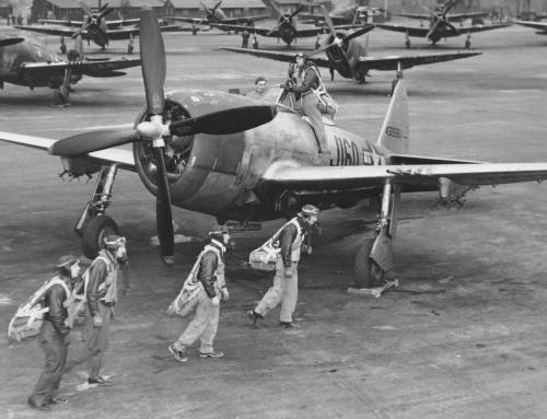 A group of pilots in flight gear walks towards a propeller-driven aircraft on a military airfield.