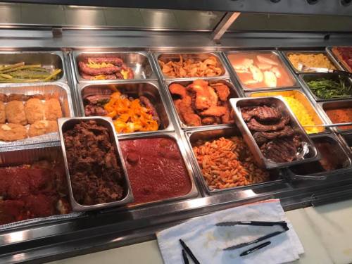 A buffet display featuring various meats, sides, and pasta in metal trays, with tongs and a napkin on the counter.