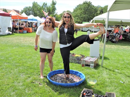 Two women joyfully standing in a blue tub filled with grapes at an outdoor event, surrounded by tents and greenery.