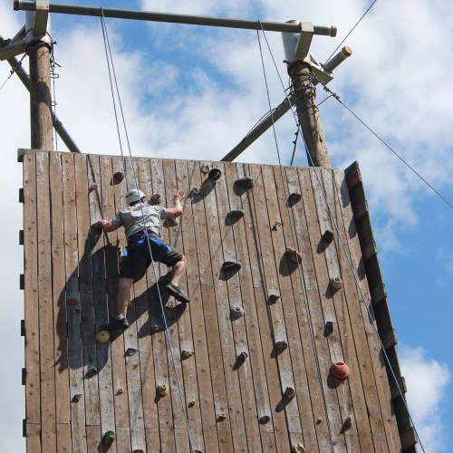 A climber ascends a wooden rock wall under a blue sky with clouds, showcasing various climbing holds.