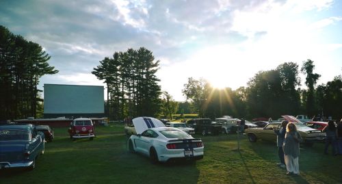 A drive-in movie scene with classic cars parked on grass, a large screen, and a sunset in the background.