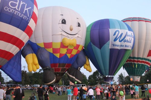 Colorful hot air balloons, including a large clown balloon, fill the sky as crowds gather below at a festival.