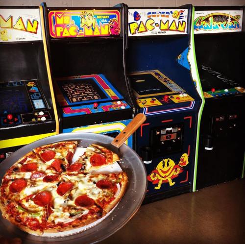 A pizza with pepperoni is held in front of classic arcade game machines, including Pac-Man and Galaga.
