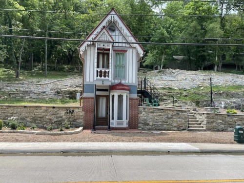A unique, narrow house with decorative features, set against a green hillside and stone landscaping.