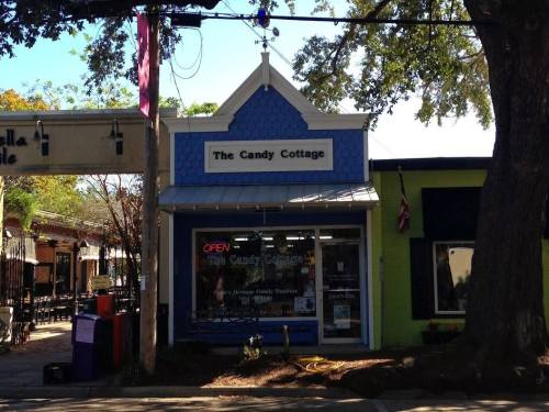 A colorful storefront of "The Candy Cottage" with an open sign, surrounded by trees and other shops.