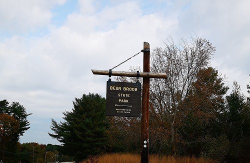 Sign for Bear Brook State Park in New Hampshire, mounted on a wooden post, surrounded by trees and cloudy skies.