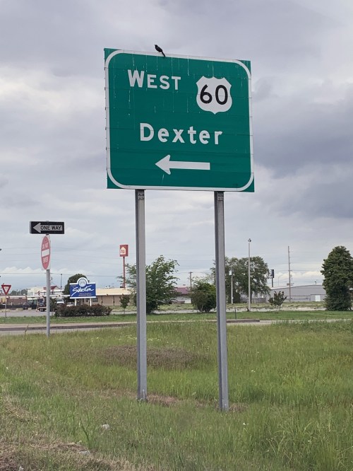 Green highway sign indicating "West 60" with an arrow pointing left towards Dexter. Cloudy sky in the background.