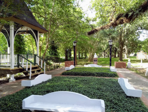 A peaceful park scene featuring a gazebo, manicured greenery, and decorative lamps under lush trees.