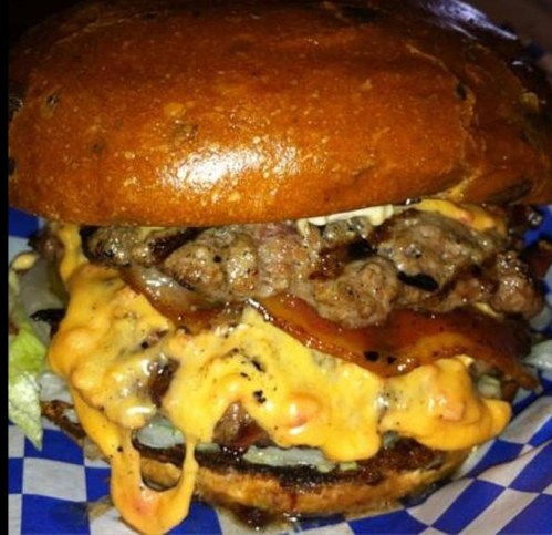 A close-up of a juicy burger with melted cheese, bacon, lettuce, and a toasted bun on a blue checkered plate.