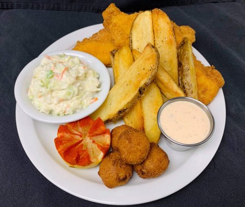 A plate of fried fish, golden potato wedges, coleslaw, a slice of tomato, and hushpuppies with dipping sauce.