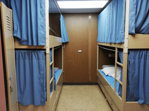 A narrow room with two sets of bunk beds, each with blue curtains and light blue bedding, against wooden walls.