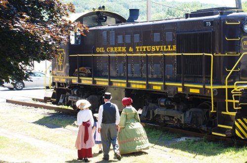 A vintage train with "Oil Creek & Titusville" on the side, alongside people in historical costumes.