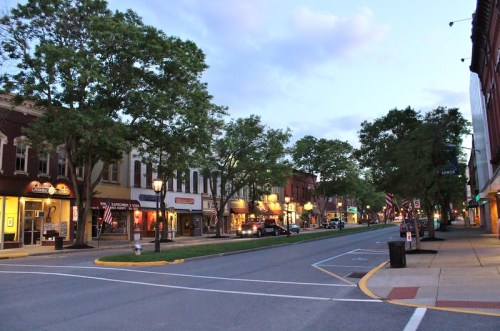 Quiet street scene at dusk, featuring shops, trees, and American flags lining the road.