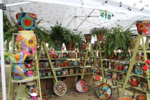 Colorful painted pots and planters displayed on wooden shelves under a white tent, surrounded by greenery.