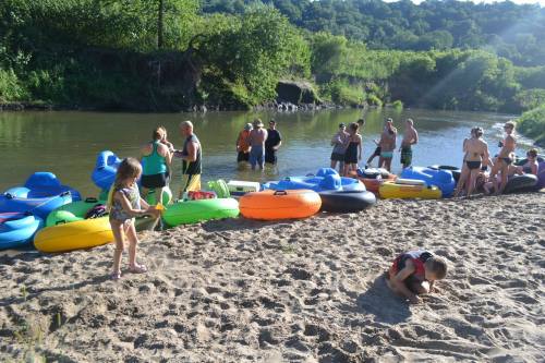 A sandy riverbank with colorful kayaks and people preparing for a water activity on a sunny day.
