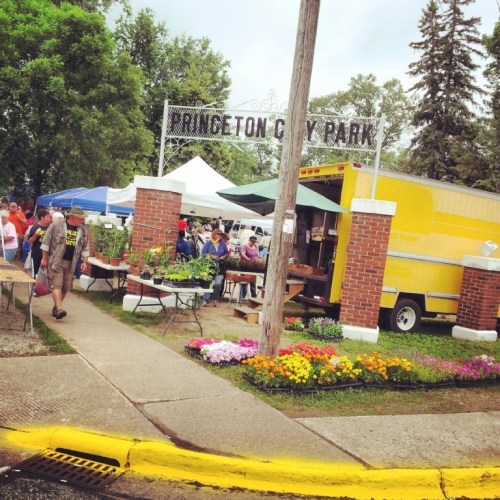 A vibrant farmers market at Princeton City Park, featuring stalls with plants and flowers, and a food truck nearby.