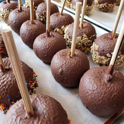 A close-up of chocolate-covered caramel apples with wooden sticks, some decorated with sprinkles and nuts.