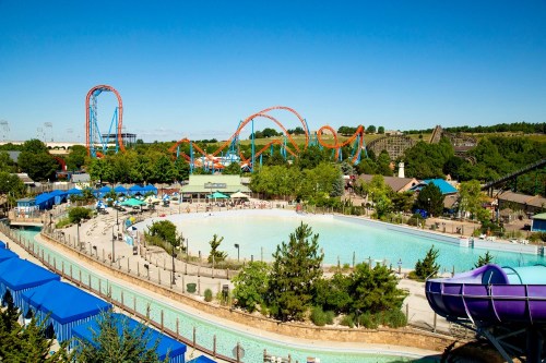 A vibrant amusement park scene featuring roller coasters, a water area, and lush greenery under a clear blue sky.