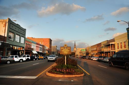 A street view of a small town with historic buildings, a sign, and parked cars under a blue sky with clouds.