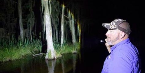 A man in a cap stands by a dark waterway, shining a flashlight on trees in a nighttime setting.