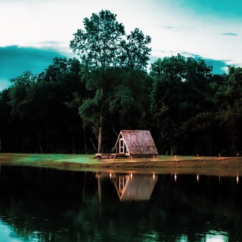A small cabin by a calm lake, surrounded by trees, reflecting the evening sky.