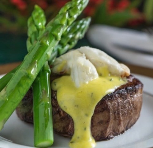 A plated dish featuring a steak topped with hollandaise sauce and crab, accompanied by asparagus spears.