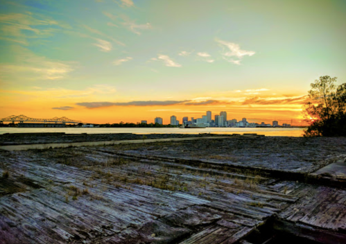 Sunset over a city skyline with a river in the foreground and wooden docks, creating a serene, picturesque scene.