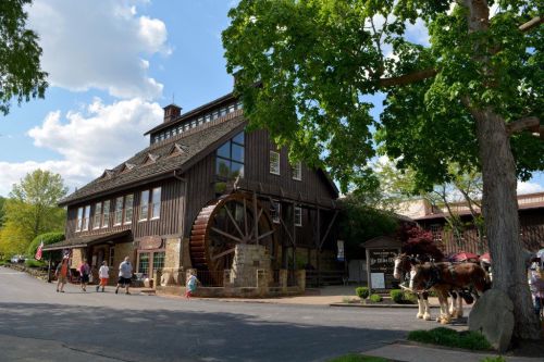 A large wooden building with a water wheel, surrounded by trees and people walking, on a sunny day.