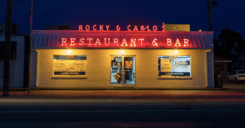 Neon-lit exterior of Rocky & Carlo's Restaurant and Bar at night, featuring inviting windows and a classic sign.