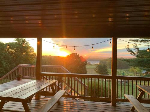 Sunset view from a wooden deck, with string lights and a scenic landscape of trees and fields in the background.