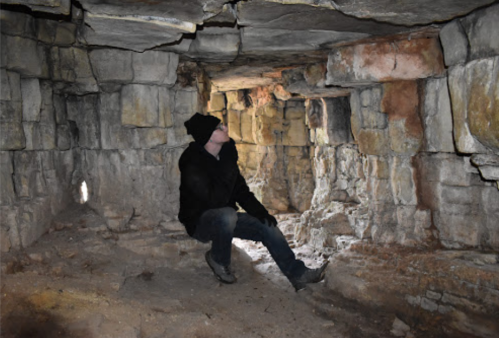 A person in a black coat and hat sits on the ground inside a stone cave, surrounded by rough, ancient walls.