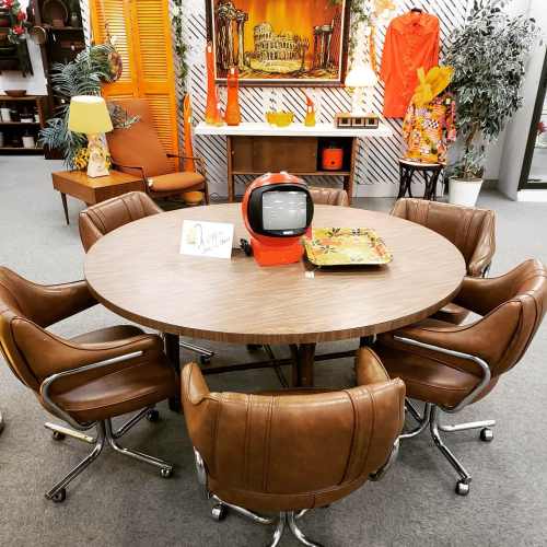 A retro dining set with a round table and six brown swivel chairs, featuring an orange vintage TV as a centerpiece.