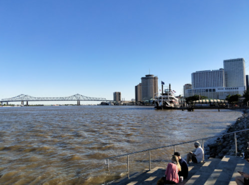 View of a river with a bridge, city skyline, and people sitting on steps by the water on a clear day.