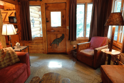 Cozy log cabin interior with a wooden door, two armchairs, a lamp, and large windows letting in natural light.