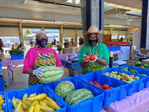 Two women in colorful outfits and masks stand behind tables filled with fresh produce at a market.