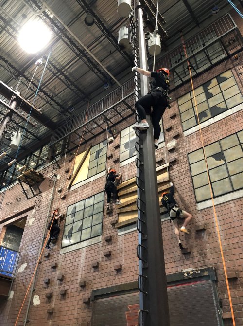 Four individuals in climbing gear ascend a tall pole inside an industrial training facility.