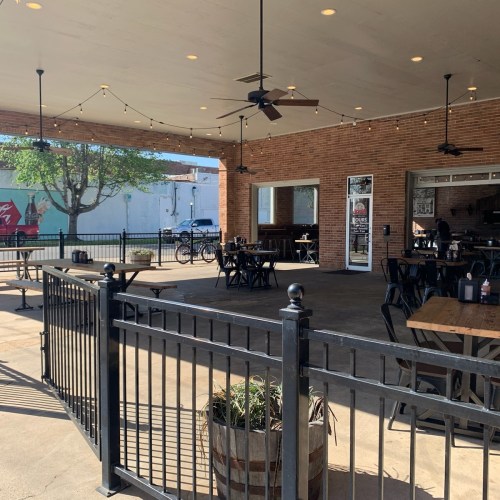 Outdoor dining area with wooden tables, string lights, and a brick wall, surrounded by a black fence.