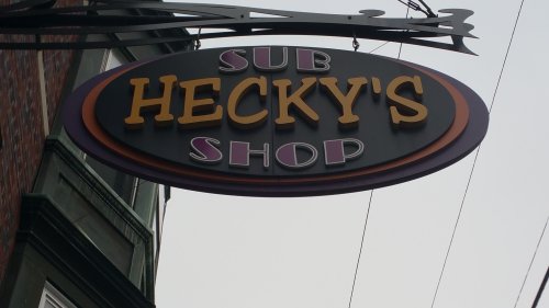 Sign for "Hecky's Shop" featuring the word "Sub" above the shop name, hanging from a building.